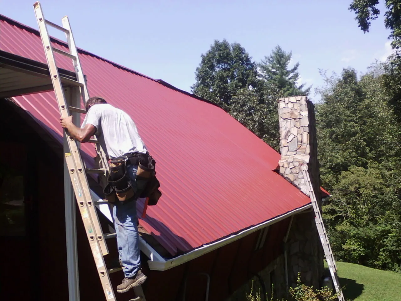 Roofer on a ladder beside a red metal roof and stone chimney — Soffit & Fascia Repair by Roofing Brothers in Rising City