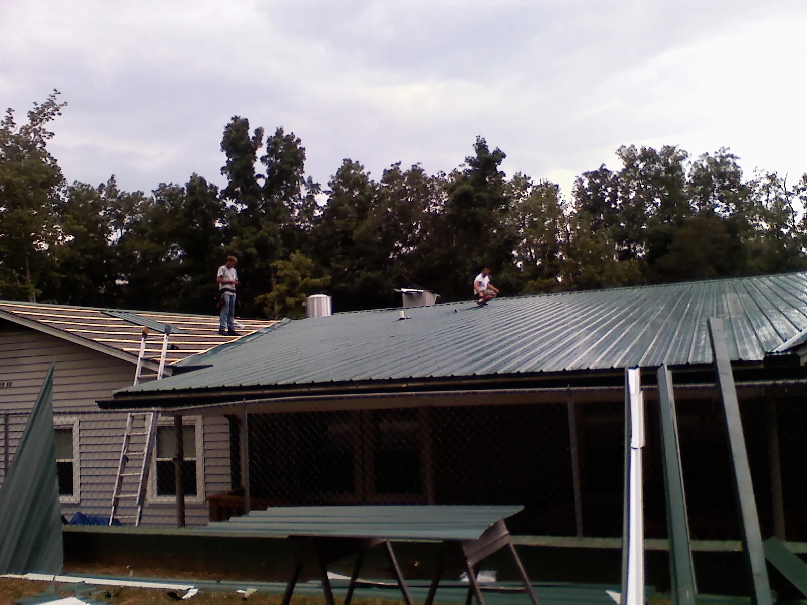 Two roofers installing green metal panels above a covered porch in Rising City